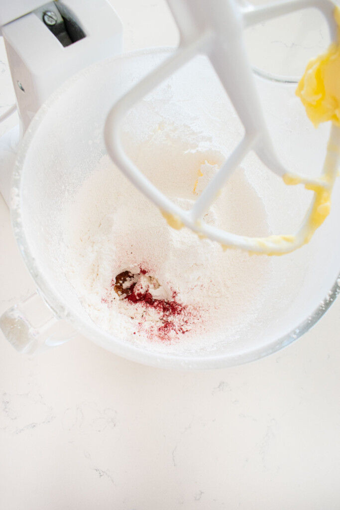 ingredients for pink vanilla frosting in a glass stand mixing bowl on a white marble counter.