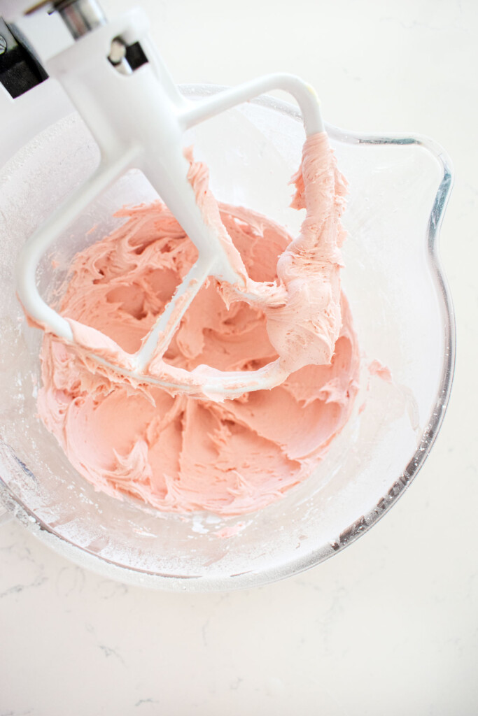 pink vanilla frosting in a glass stand mixing bowl on a white marble counter.