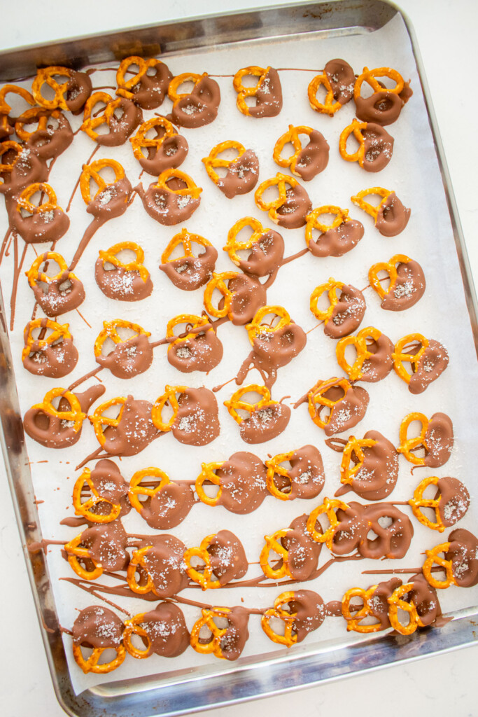 mini pretzel twists dipped in milk chocolate with sea salt on a baking sheet with parchment paper on a marble counter.