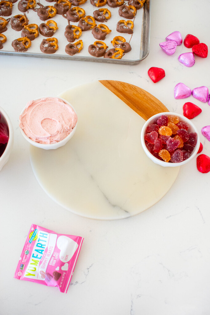 charcuterie board with a bowl of pink frosting and a bowl of heart gummies on a white marble counter.