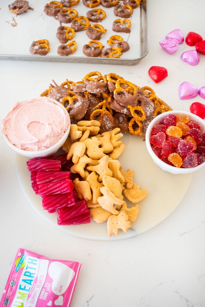 charcuterie board with a bowl of pink frosting, a bowl of heart gummies, licorice, chocolate pretzels and animal crackers on a white marble counter.