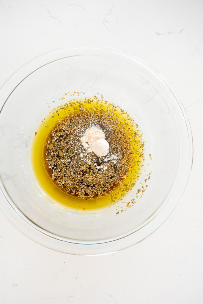 ingredients for greek salad dressing in a glass bowl on a white marble counter.