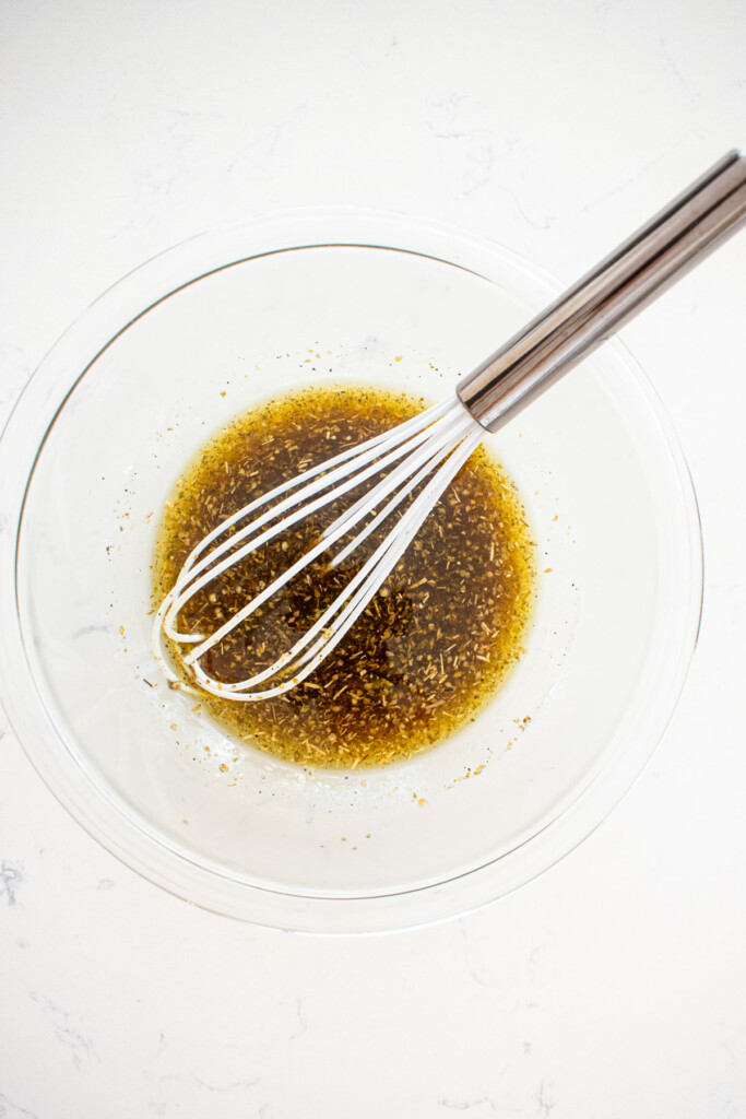 greek salad dressing mixed in a glass mixing bowl with a whisk on a white marble counter.