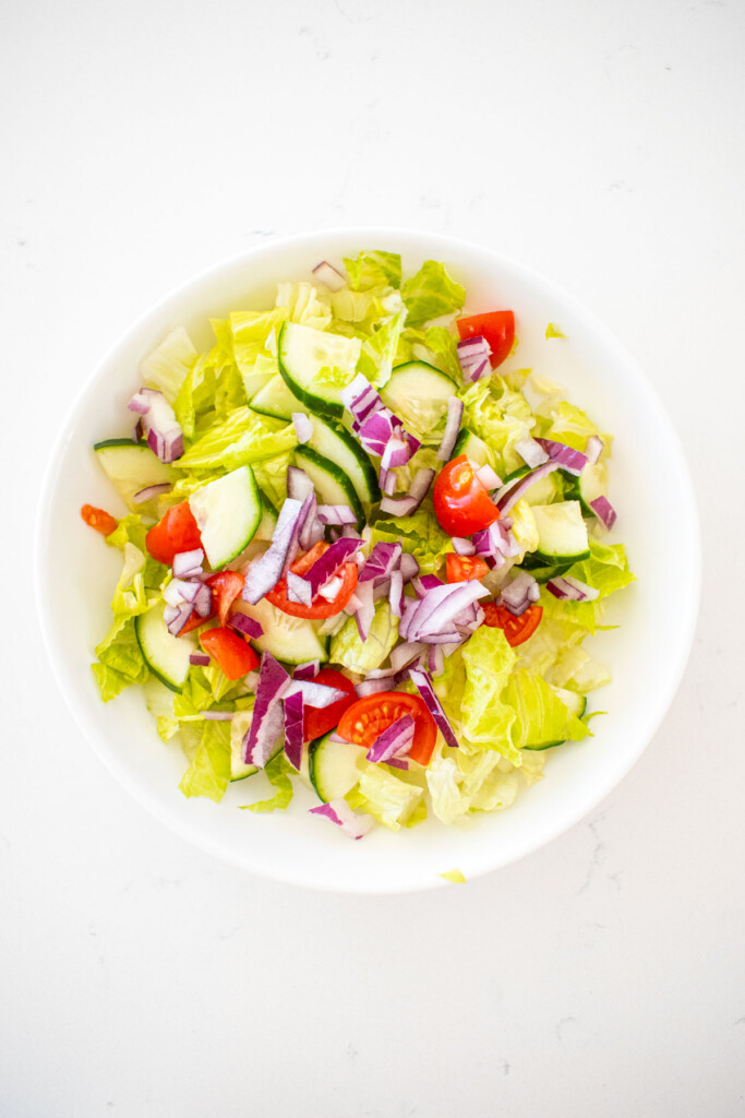 romaine lettuce, cucumber, red onion and tomato in a big white salad bowl on a marble counter.