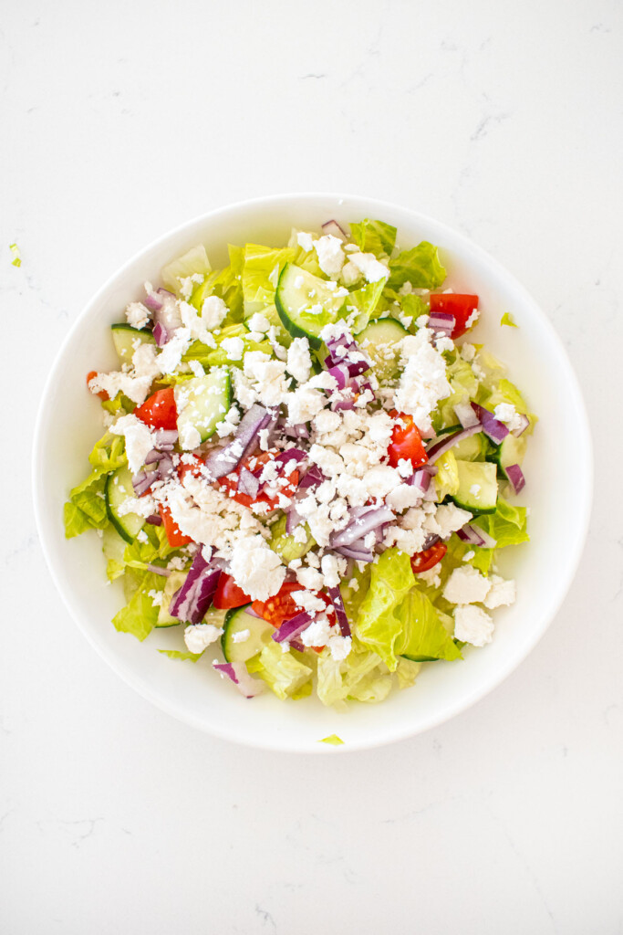 romaine lettuce, cucumber, red onion, tomato and feta in a big white salad bowl on a marble counter.