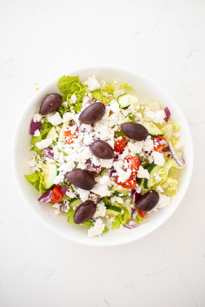 romaine lettuce, cucumber, red onion, tomato, feta, and kalamata olives in a big white salad bowl on a marble counter.