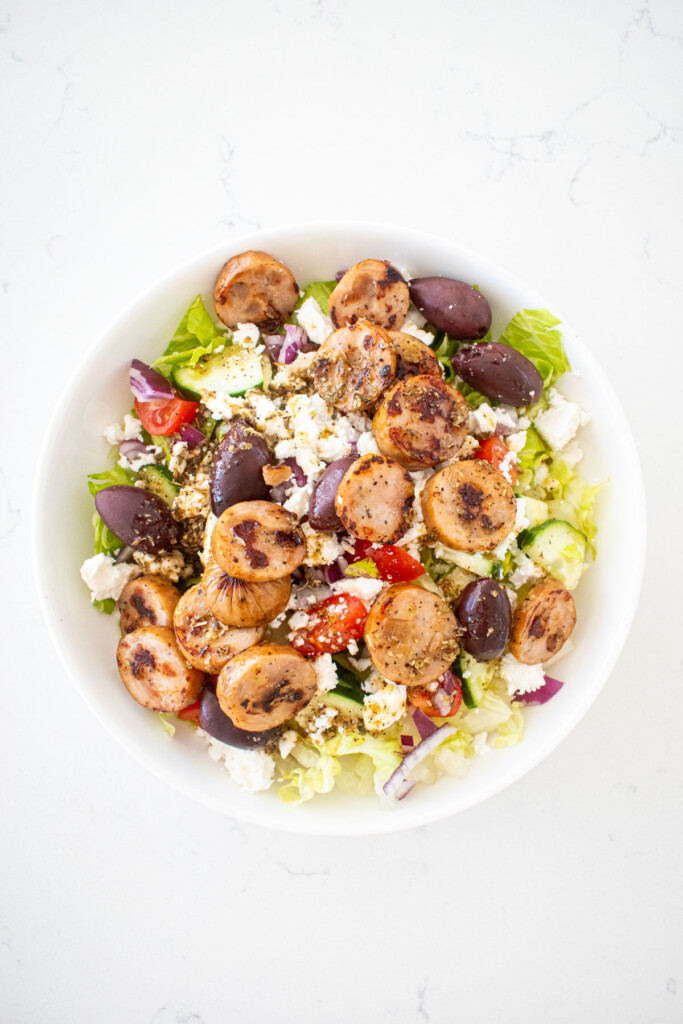 chicken sausage greek salad with salad dressing in a bowl on a white marble counter.