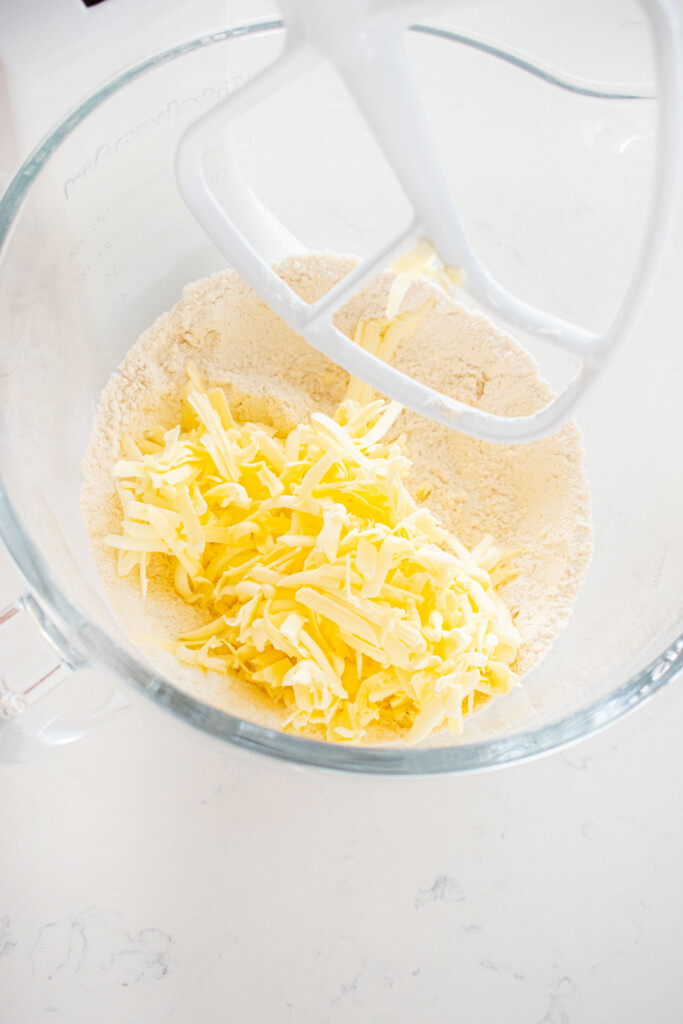 grated butter on top of dry ingredients in a glass mixing bowl on a white marble counter.