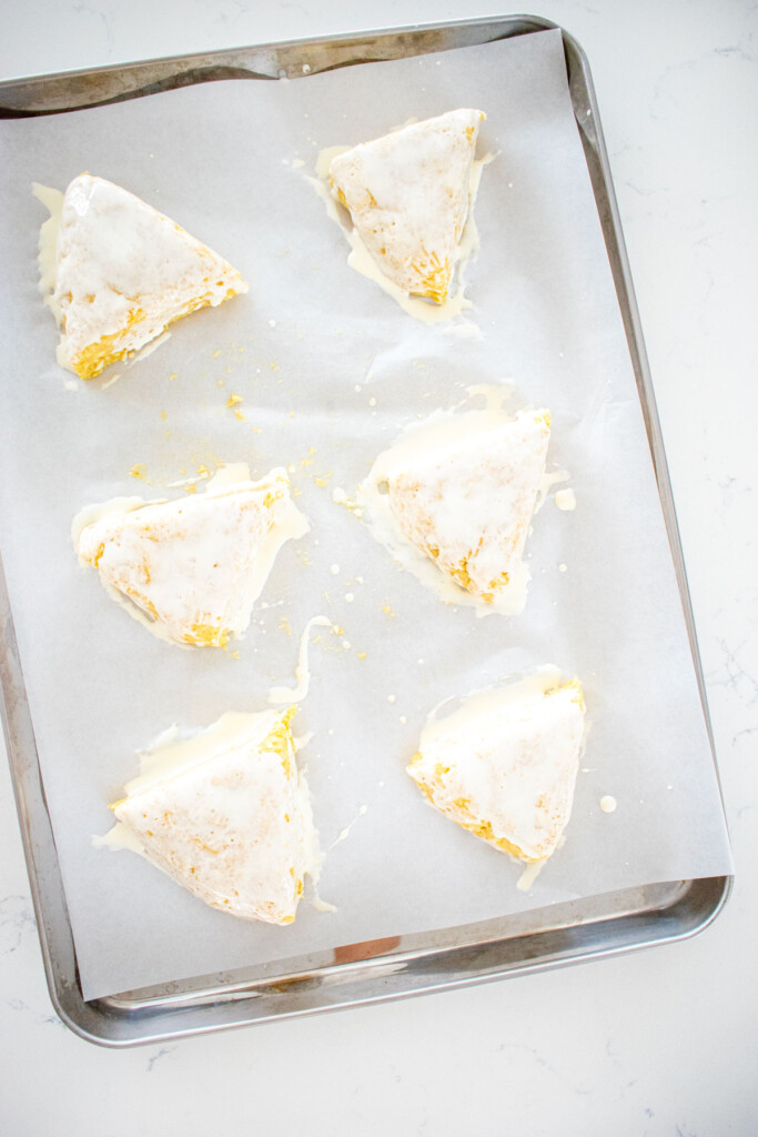 citrus scones dough cut into 6 triangles spread out evenly on a baking sheet with cream brushed on top on a white marble counter.