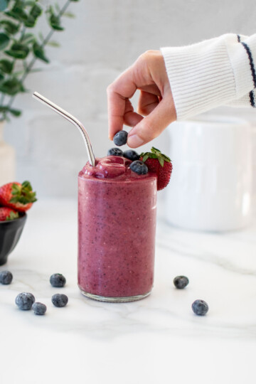 woman placing a blueberry on top of a berry protein smoothie on a marble countertop.