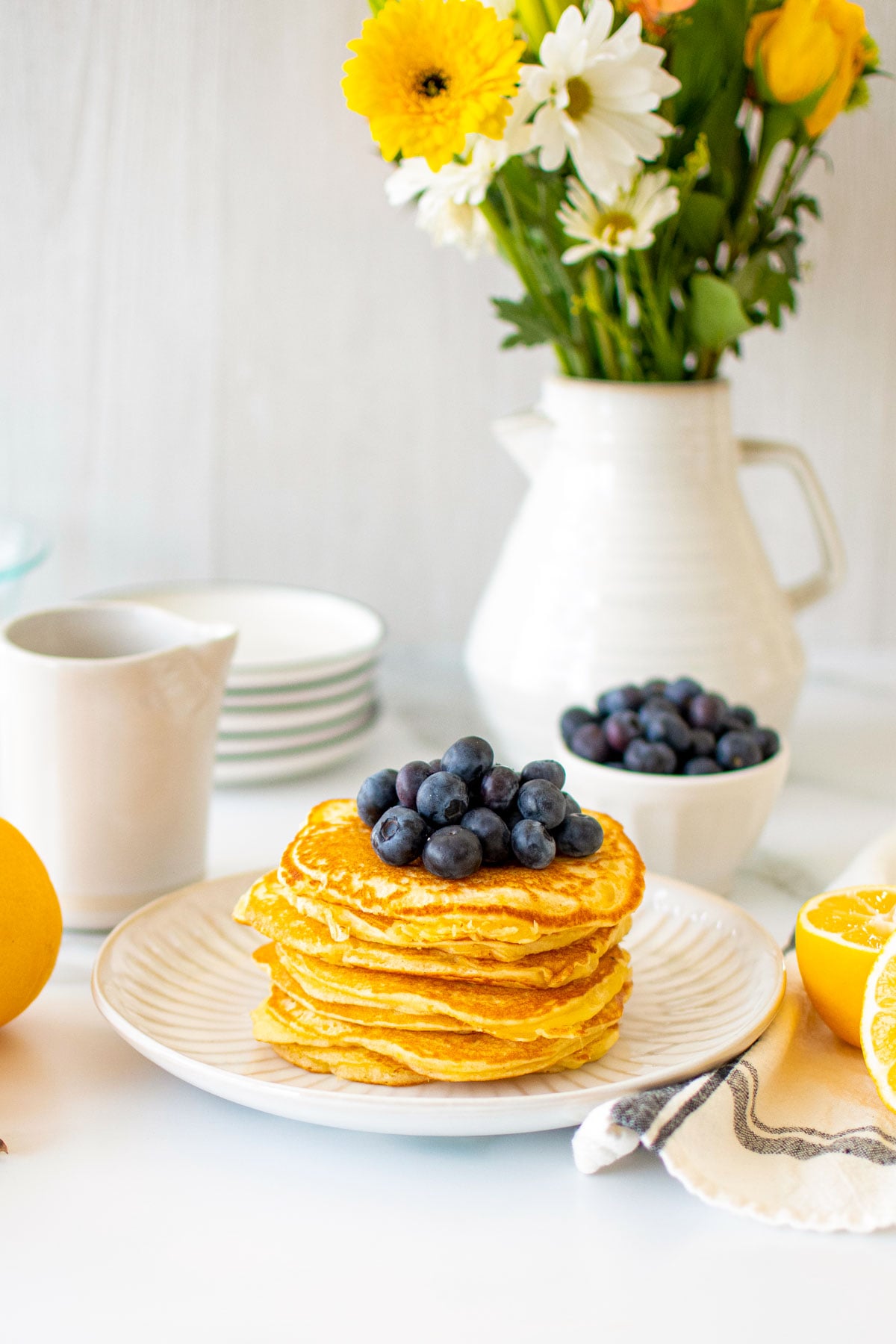 stack of meyer lemon pancakes on a plate with fresh blueberries on a white marble counter with fresh flowers.