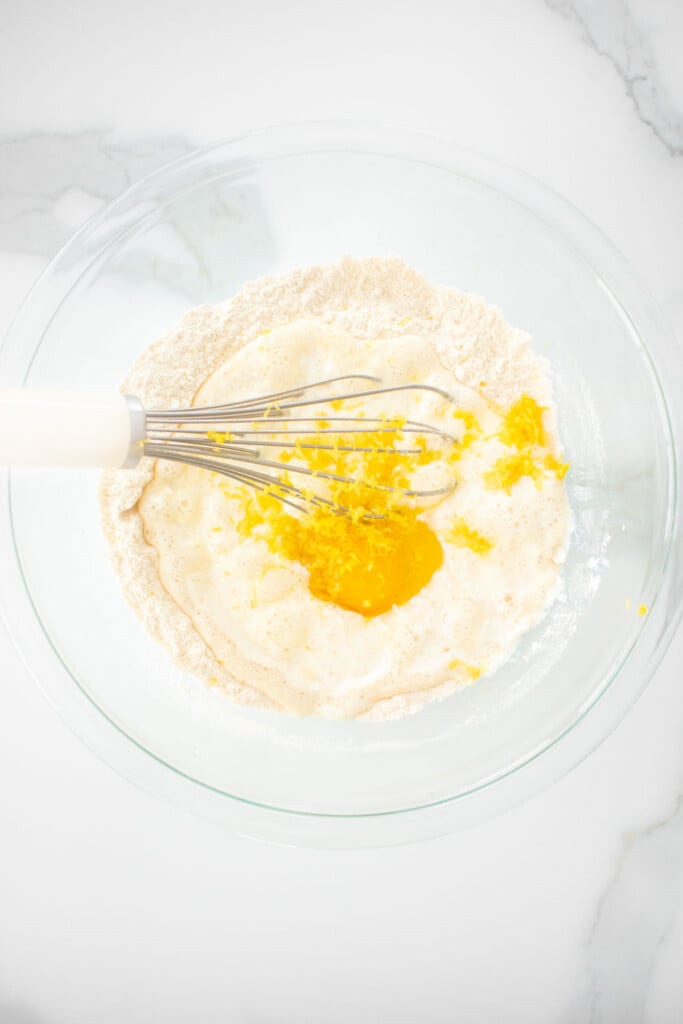 eggs, milk and lemon zest added into dry ingredients for meyer lemon pancakes in a glass mixing bowl with a whisk.