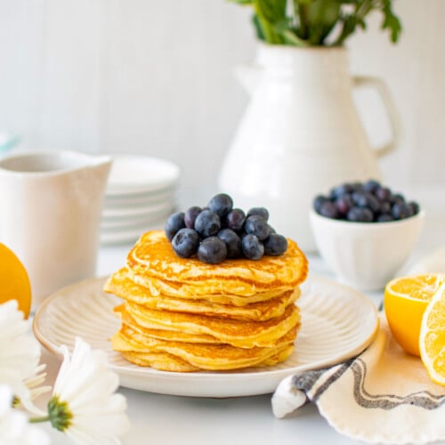 stack of meyer lemon pancakes on a plate with fresh blueberries on a white marble counter with fresh flowers.