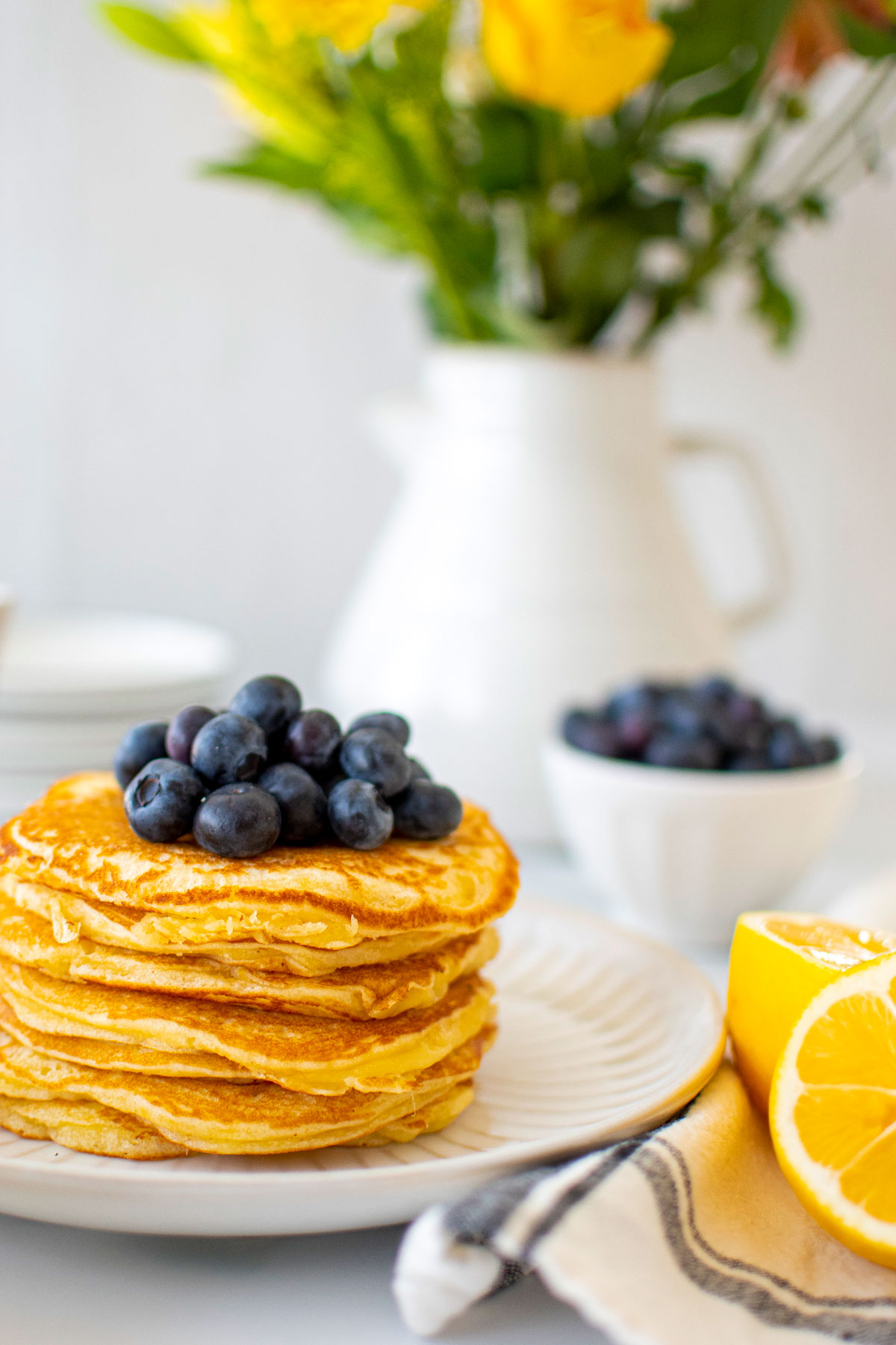 stack of meyer lemon pancakes on a plate with fresh blueberries on a white marble counter with fresh flowers.