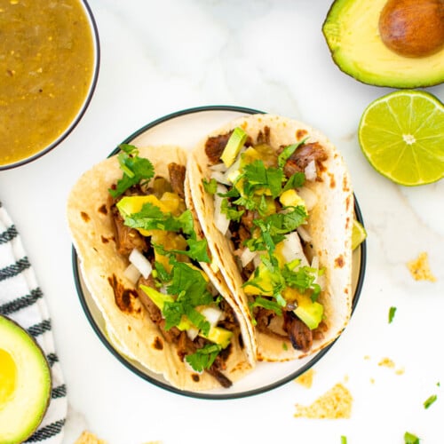 slow cooker pulled pork street tacos on a plate with chips and avocado on a marble countertop.