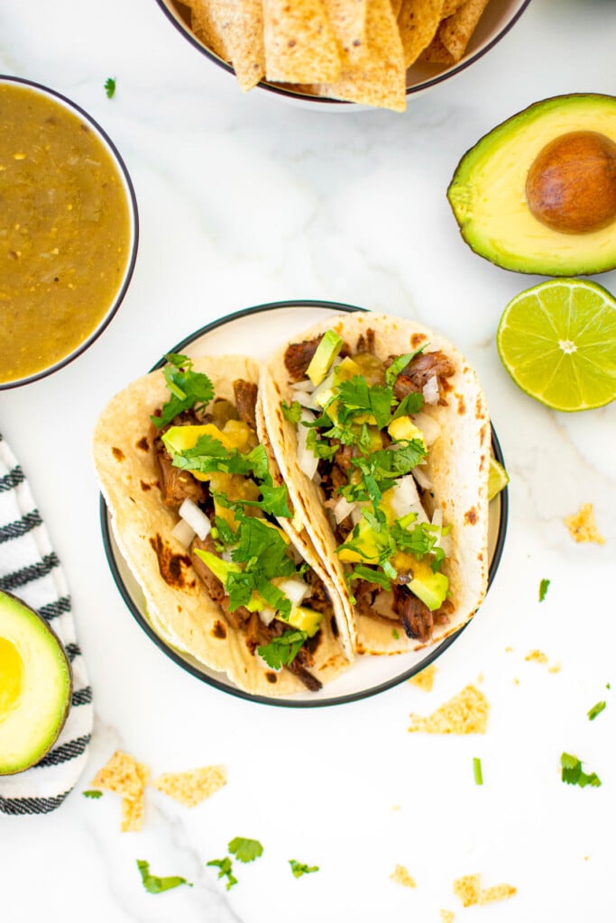 slow cooker pulled pork street tacos on a plate with chips and avocado on a marble countertop.