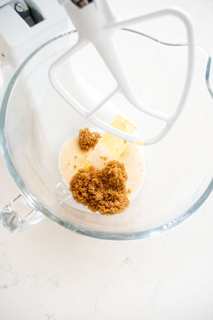 butter, sugar and brown sugar in a glass stand mixing bowl on a white marble counte.r