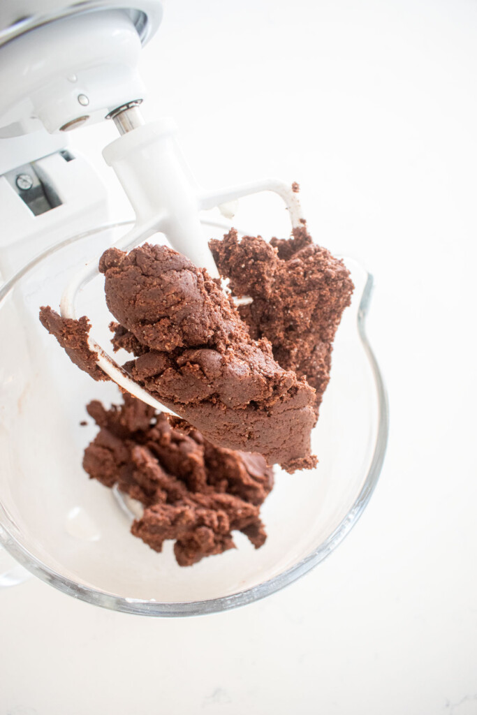 chocolate biscotti dough in a glass stand mixing bowl on a white marble counter.