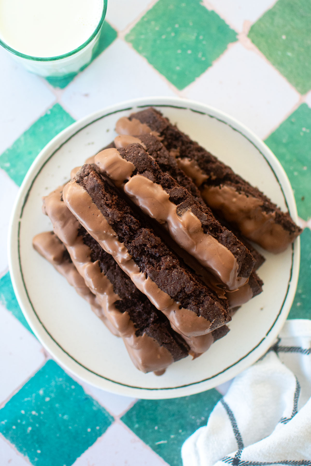 thin mint biscotti dipped in milk chocolate on a plate with a glass of milk on a green and white tile table.