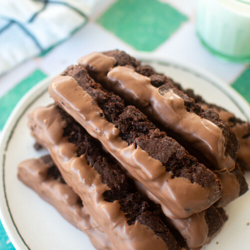 thin mint biscotti dipped in milk chocolate on a plate with a glass of milk on a green and white tile table.