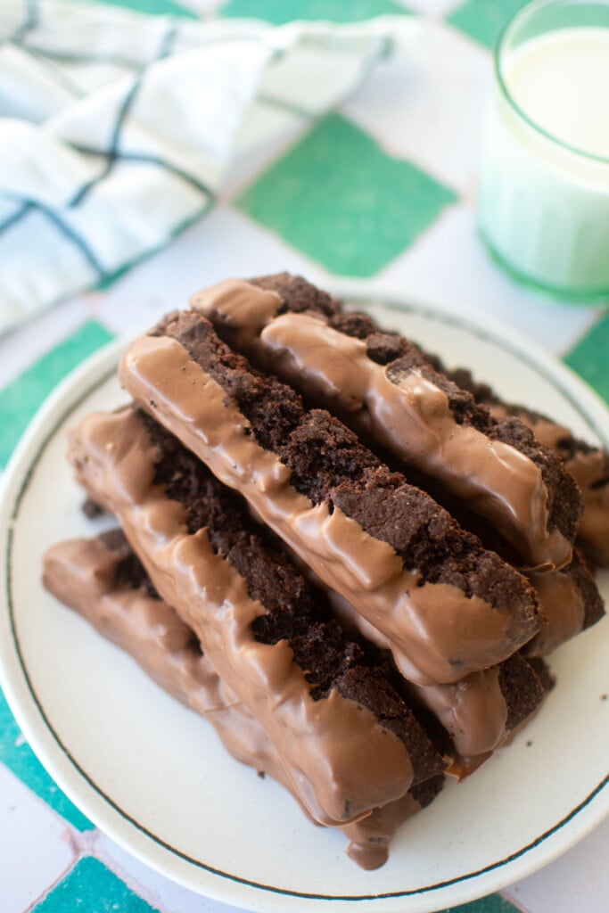 thin mint biscotti dipped in milk chocolate on a plate with a glass of milk on a green and white tile table.