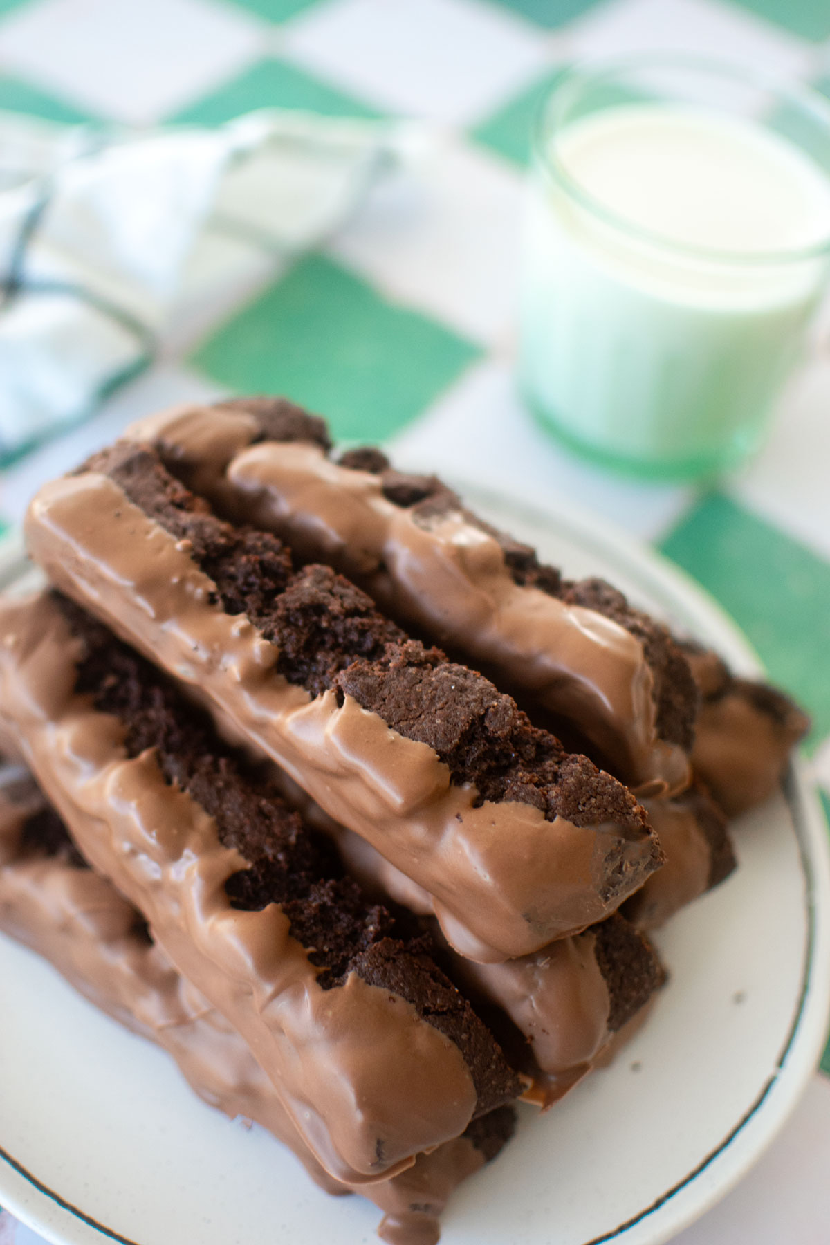 thin mint biscotti dipped in milk chocolate on a plate with a glass of milk on a green and white tile table.