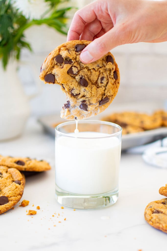 vegan banana chocolate chip cookie being dunked into a glass of milk on a marble counter.