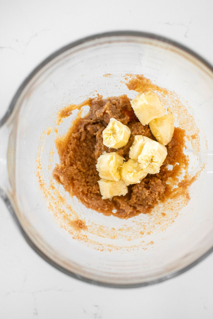 creamed coconut oil and sugar in a glass mixing bowl with banana chunks on a marble counter.