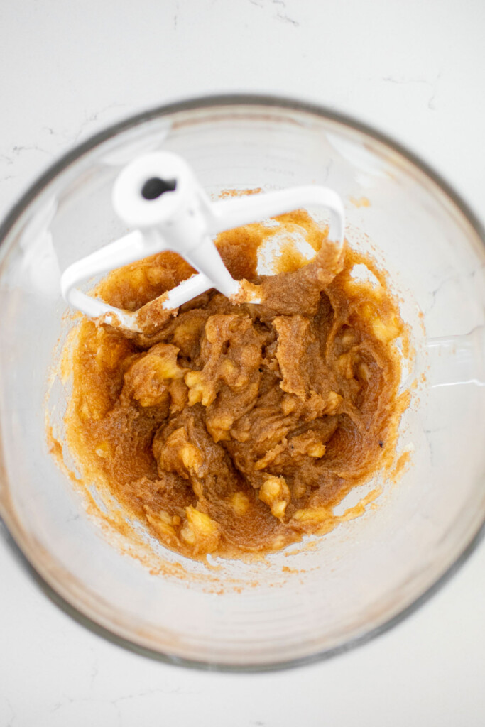 creamed coconut oil, sugar and banana in a glass mixing bowl with banana chunks on a marble counter.