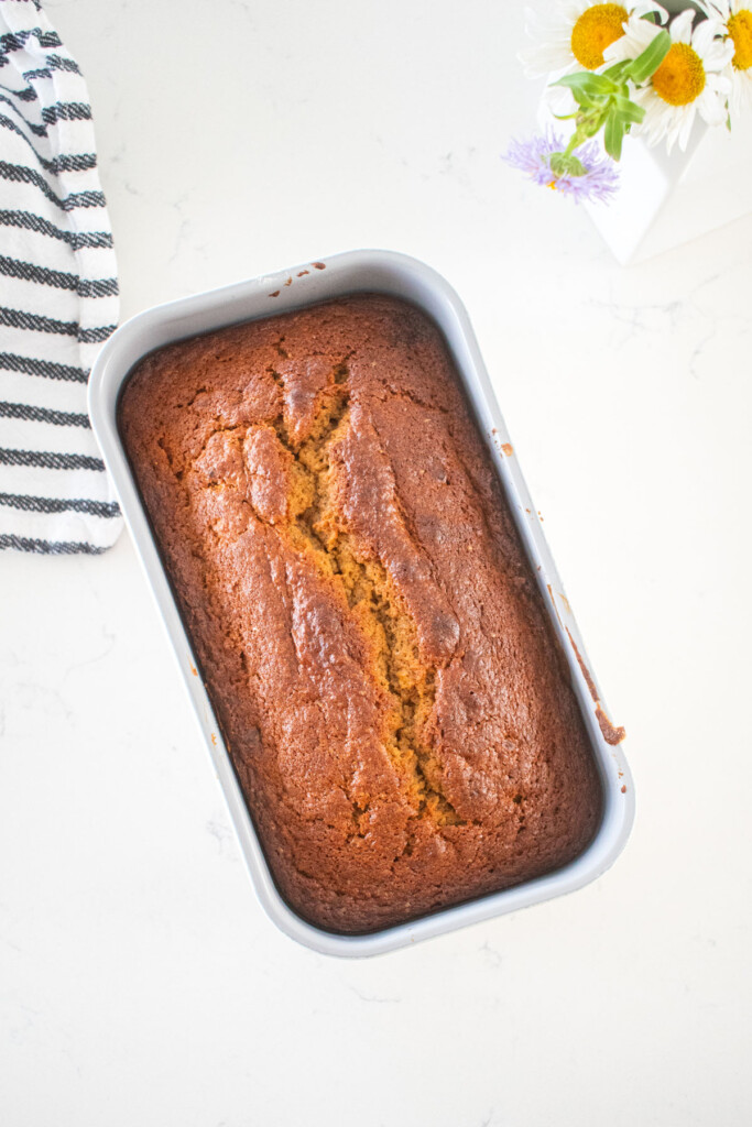 baked almond butter banana bread in a loaf pan on a marble counter with fresh flowers.