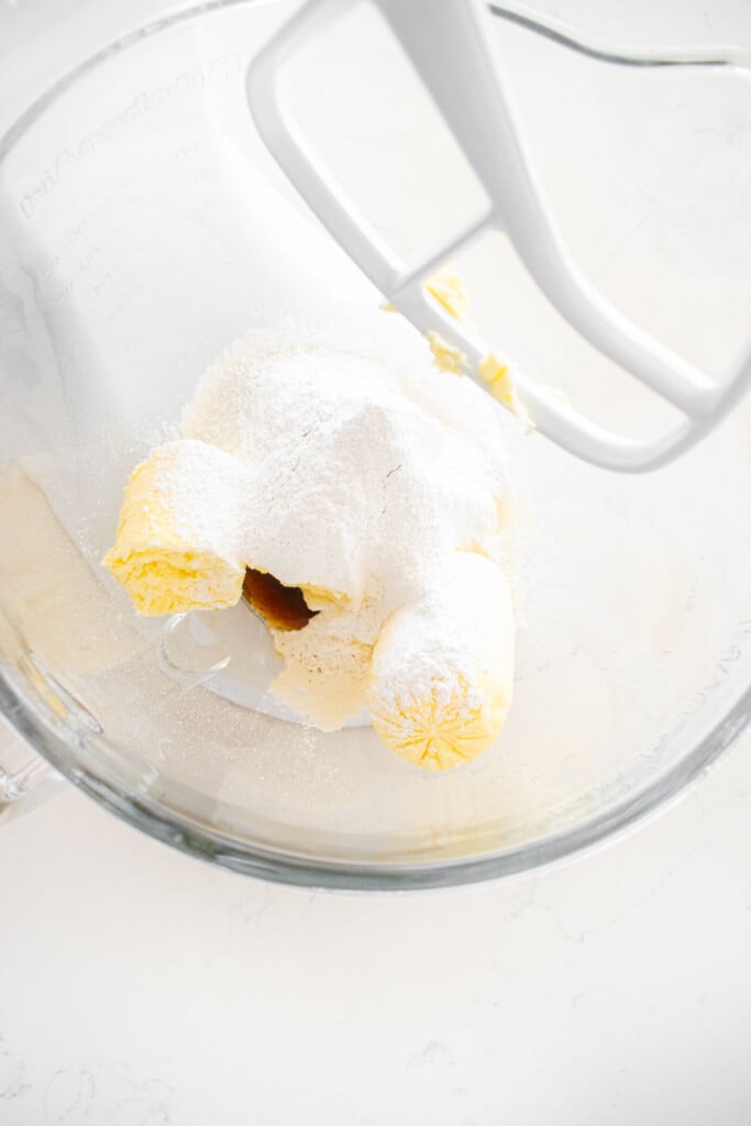glass stand mixing bowl with butter, powdered sugar and vanilla extract on a white marble counter.