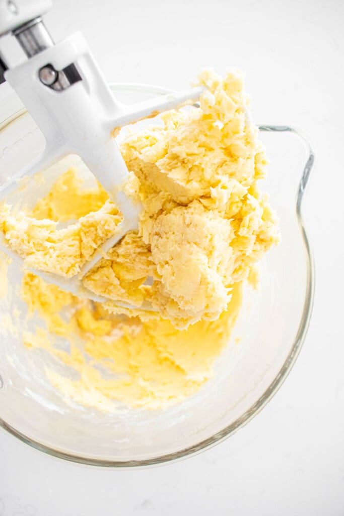 glass stand mixing bowl with creamed butter, powdered sugar and vanilla extract on a white marble counter.