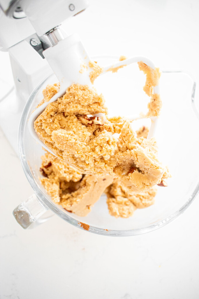 pecans mixed into cinnamon pecan cookie dough in a glass stand mixing bowl on a white marble counter.