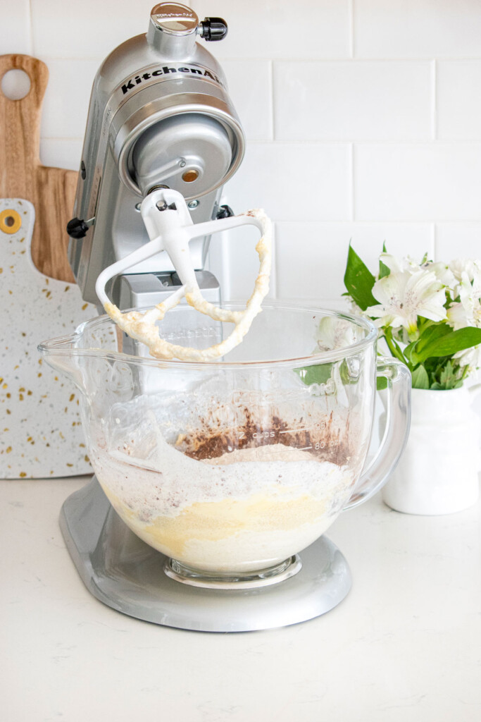 ingredients for eggless chocolate cake in a glass stand mixing bowl on a white marble counter with fresh flowers.