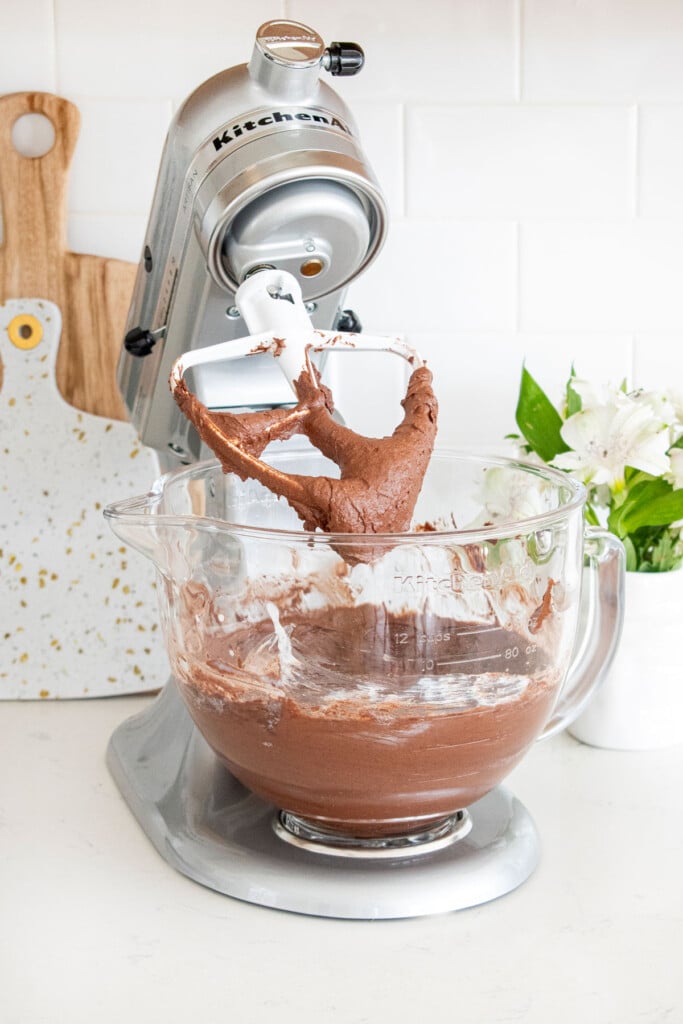 eggless chocolate cake batter in a glass stand mixing bowl on a white marble counter.