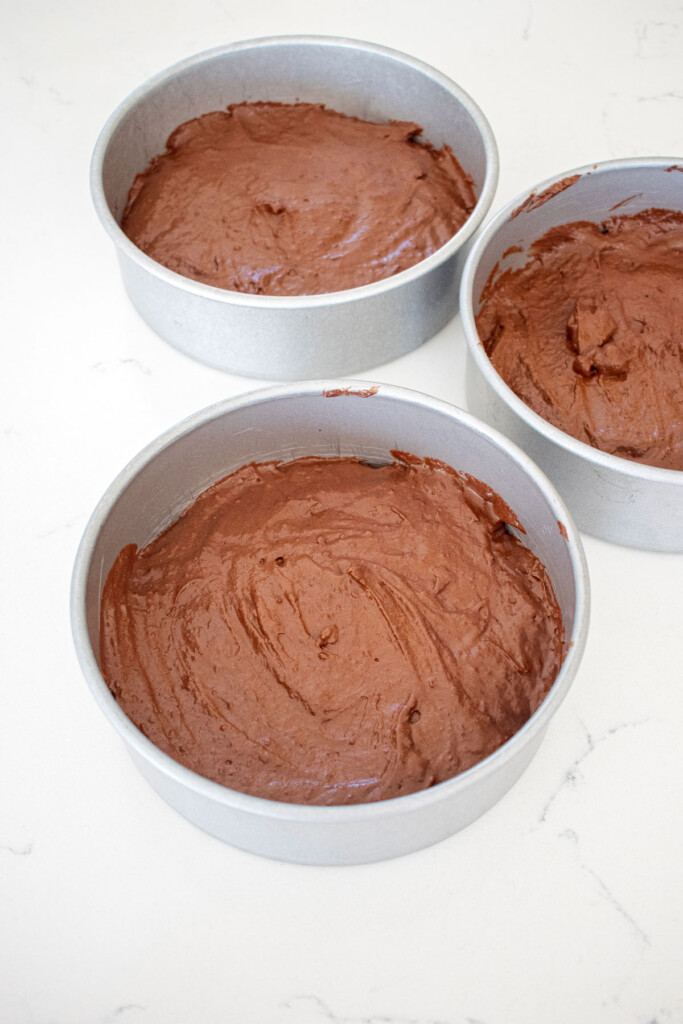 eggless chocolate cake batter in three cake pans on a white marble counter.
