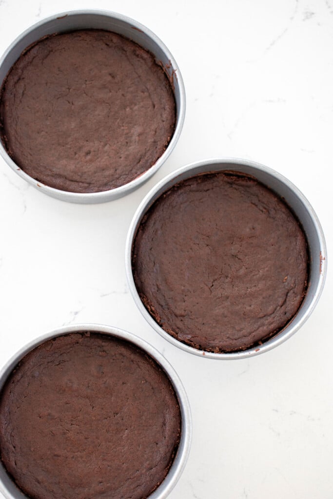chocolate cake in three cake pans sitting on a white marble counter.