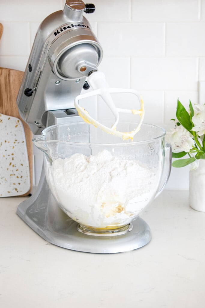 ingredients for cream cheese frosting in a glass stand mixing bowl on a white marble counter with fresh flowers.