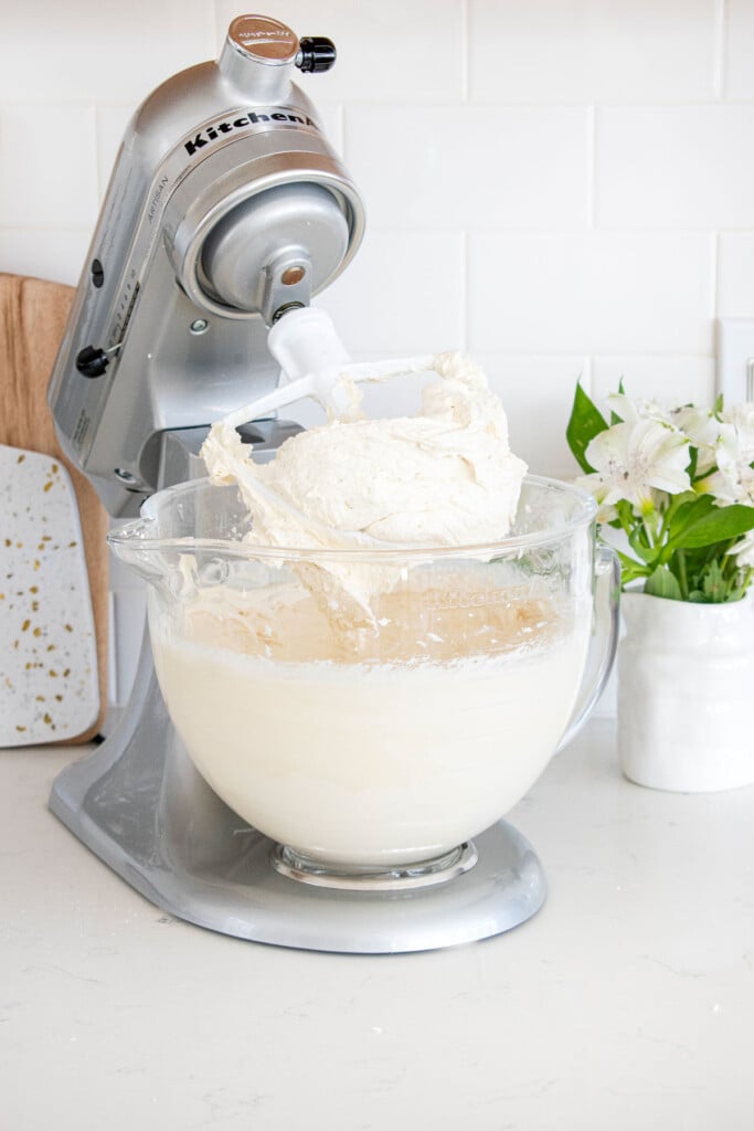 cream cheese frosting in a glass stand mixing bowl on a white marble counter with fresh flowers.