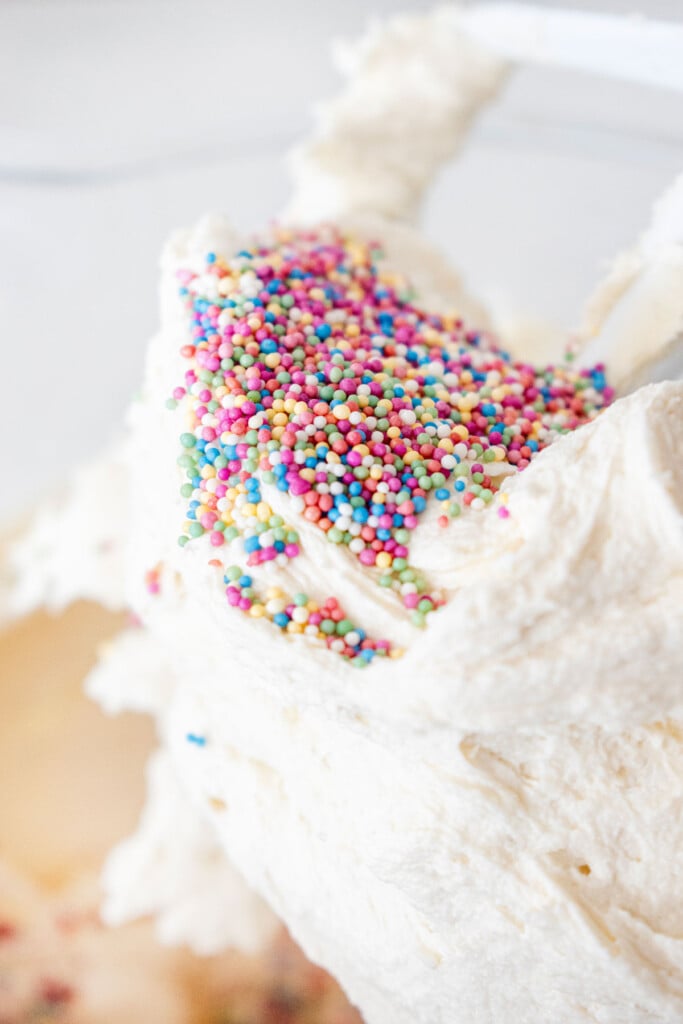 nonpareils added to cream cheese frosting in a glass bowl of a stand mixer on a white marble counter.