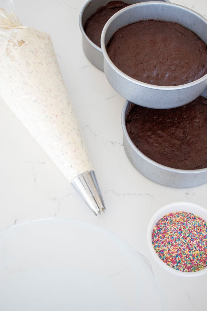 funfetti cream cheese frosting in a piping bag with a star decorating tip and chocolate cake and sprinkles on a white marble counter.