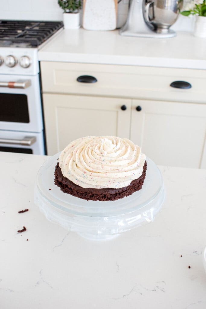 eggless birthday cake with cream cheese frosting being assembled on a cake stand on the kitchen counter.