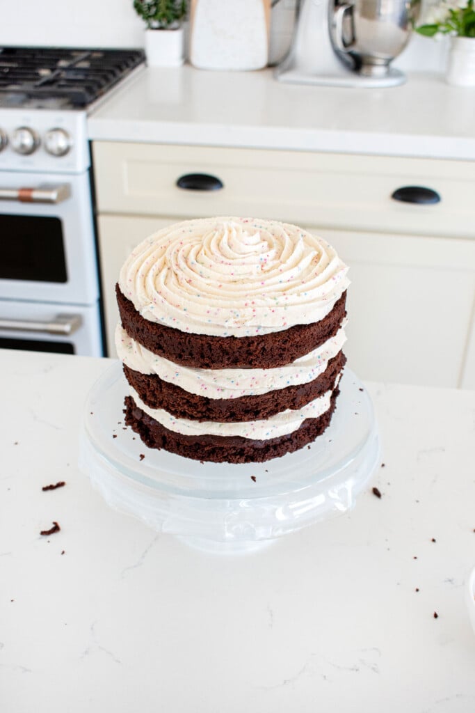 eggless birthday cake with cream cheese frosting being assembled on a cake stand on the kitchen counter.