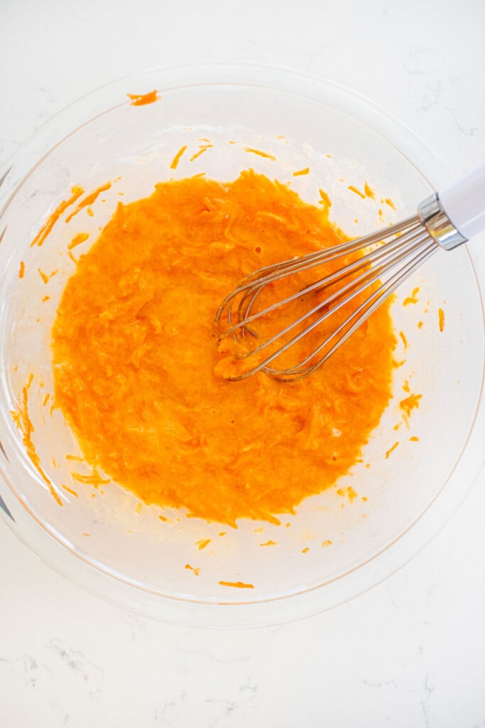 wet ingredients for orange ginger carrot cake in a glass mixing bowl with a whisk on a white marble counter.