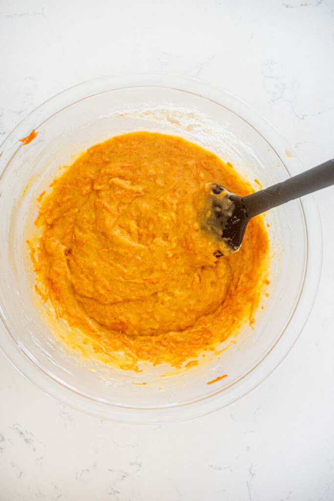 orange ginger carrot cake batter in a glass mixing bowl with a spatula on a white marble counter.