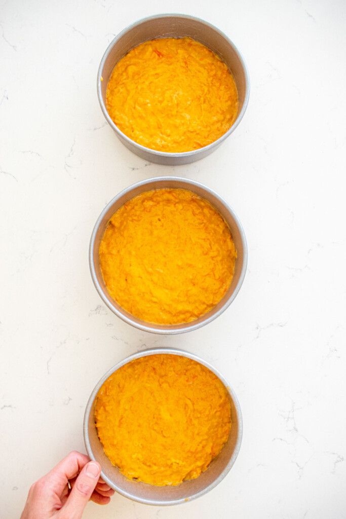 orange ginger carrot cake batter in three 6-inch cake pans on a white marble counter.
