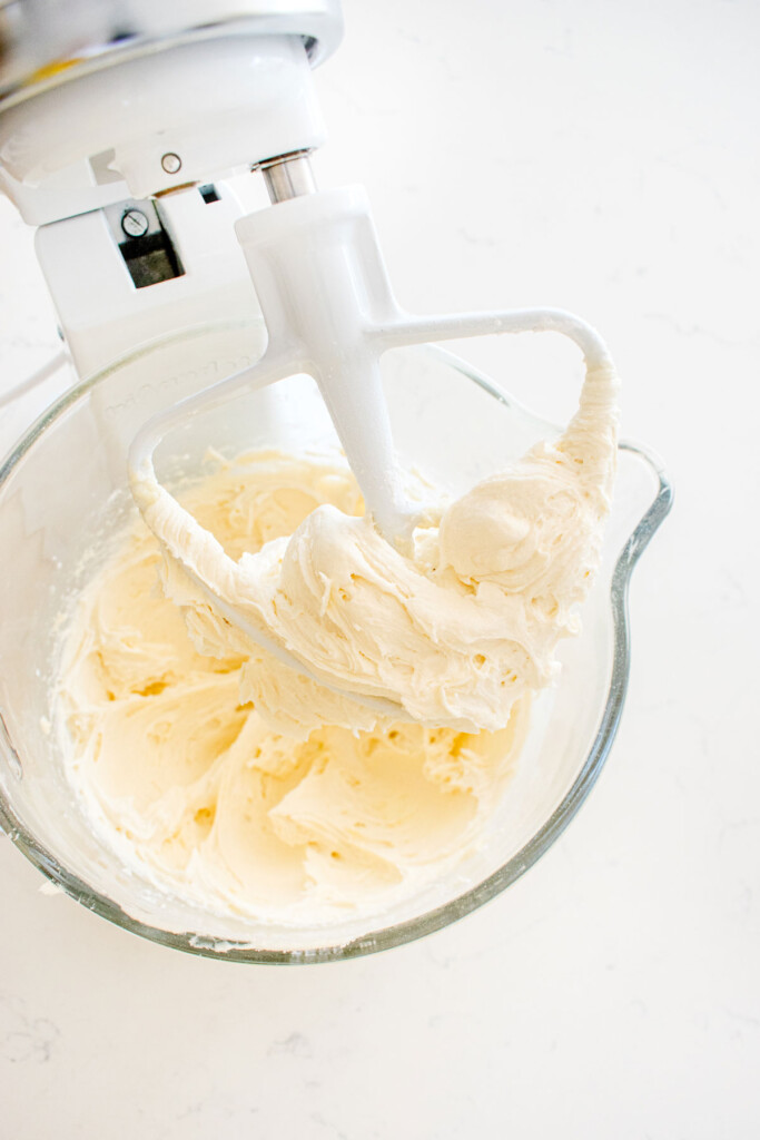 orange cream cheese frosting in a glass mixing bowl on a white marble counter.