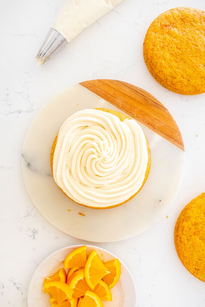 orange ginger carrot cake with a spiral of orange cream cheese buttercream frosting being assembled on a marble serving board.