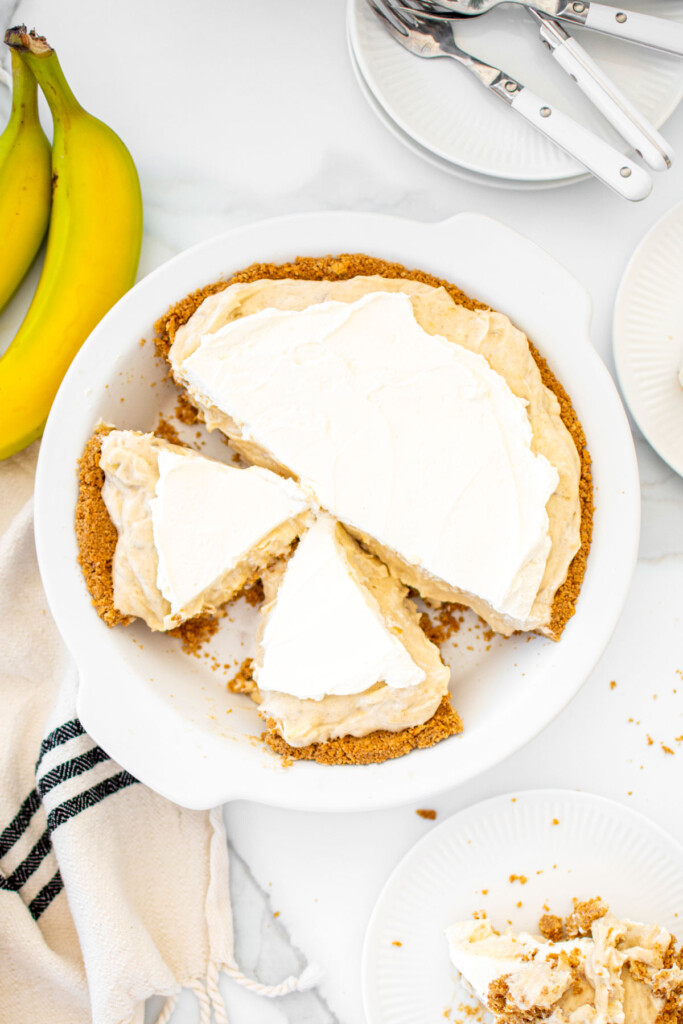 banana cream pie with graham cracker crust in a white pie pan on a white marble table cut into slices.