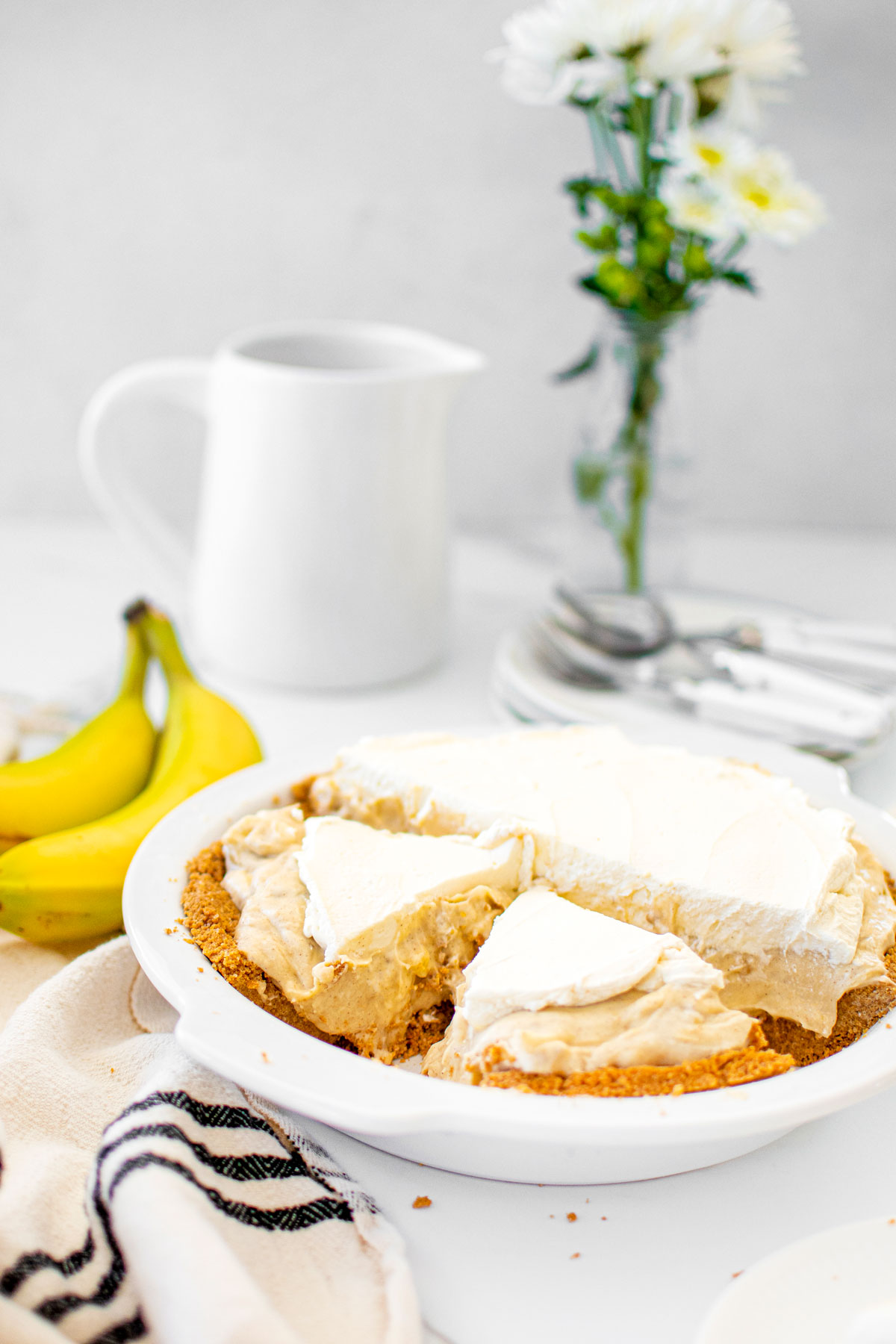 banana cream pie with graham cracker crust in a white pie pan on a white marble table cut into slices.